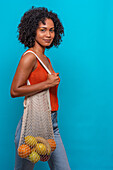 Side view of delighted African American female model with Afro hairstyle in casual clothes carrying bag of fruit on shoulder against blue background