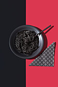 Top view of black spaghetti in a bowl with fork placed on black and red background near napkin