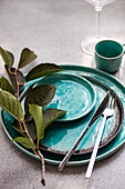 Stylish turquoise ceramic plates with matching cup, silver cutlery, and fresh cherry leaves on a textured grey surface