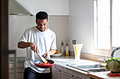 Positive young ethnic African American male in casual clothes preparing delicious dish while frying tomatoes on pan and mixing ingredients with wooden spatula during cooking process in kitchen