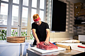 Concentrated young male with pink headband standing at table with fish and looking down while cutting fresh raw red tuna on white board near glass windows