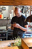 Focused bearded chef wearing black apron standing near kitchen utensils and browsing mobile phone during work in restaurant at Zermatt Switzerland