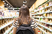 Back view of unrecognizable female customer in striped T shirt with backpack choosing products between shelves in supermarket
