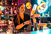 Confident beautiful young barkeeper pouring alcohol in jigger from glass bottle at bar counter while working in illuminated nightclub