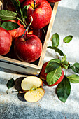 Top view of whole fresh red ripe organic delicious apples with green leaves filled in wooden box with half cut pieces and placed on gray surface in daylight