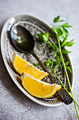 Hoher Winkel eines Löffels mit nativem Olivenöl, Zitronenscheiben und grünem Petersilienkraut auf einem Teller vor unscharfem Hintergrund