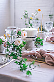 Eleganter Tisch mit Tellern und Blumen in der Nähe eines leckeren Kuchens auf einem rosa Tischtuch vor einer Backsteinwand