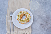 Ringförmiges Brandteiggebäck mit weißem Zuckerguss und gehackten Pistazien bestreut, präsentiert auf einem weißen Teller mit Gabel und Glas auf einem beigen Tischset auf einer grauen Oberfläche von oben