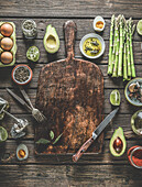Hintergrundbild mit verschiedenen gesunden Zutaten: grüner Spargel, Avocadohälften, Frühlingszwiebeln, gekochte Eierhälften, Kräuter und Gewürze auf einem hölzernen Schneidebrett mit Messer und Gabeln. Ansicht von oben