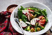 Close-up of healthy vegetable salad with fresh organic spinach leaves, juicy tomatoes, crunchy radish slices, and tangy olives served on a white plate with fork on the side all placed on textured surface with checkered cloth