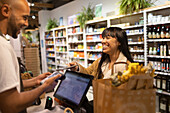 Verkäufer, afroamerikanisch, männlich, im Schurz, an einer Theke mit einem POS-Terminal, während ein asiatischer Kunde elektronisch mit einem Smartphone bezahlt