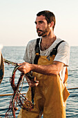 Fokussierter, bärtiger, männlicher Fischer in Uniform, der mit einem Netz auf Fischfang geht, während er auf einem Schoner in Soller nahe der Baleareninsel Mallorca arbeitet