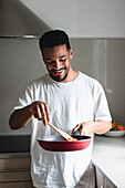 Positive young ethnic African American male in casual clothes preparing delicious dish while frying tomatoes on pan and mixing ingredients with wooden spatula during cooking process in kitchen