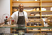 Cheerful young African American male seller in apron smiling and looking at camera while standing behind counter against shelves