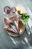 Draufsicht auf einen elegant gedeckten Frühlingstisch mit gestapelten Keramiktellern, Leinenservietten, Besteck und Gläsern, begleitet von zarten Rosen auf einer strukturierten Oberfläche