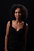 Smiling young Hispanic female model with Afro hairstyle in black clothes looking at camera while standing against black background