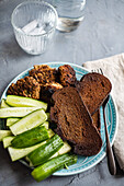 High angle of fresh organic cucumber, slices of rye sour dough bread and meat cutlets served on plate near jar of water, glass, fork and napkin against gray background