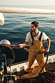 Von oben fokussierter bärtiger männlicher Fischer in Uniform, der mit einem Netz auf Fischfang geht, während er auf einem Schoner in Soller nahe der Baleareninsel Mallorca arbeitet