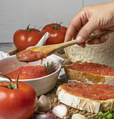 Crop hand of anonymous cook taking tomato spread from bowl with wooden spoon for applying on bread near garlic on table