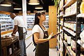 Side view of African American female sales staff in apron standing near shelves with assorted goods and checking with help of tablet while working