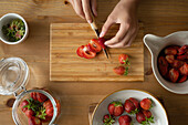 From above of anonymous person with knife slicing strawberries on wooden board and during strawberry jam preparation indoors