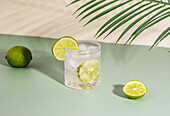 Blick von oben auf ein funkelndes, transparentes Kristallglas mit einem erfrischenden Cocktail mit Zitrusscheiben auf einem Tisch mit Limetten und einem halbierten Stück neben grünen Blättern