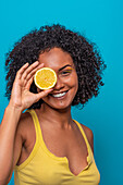 Happy young Hispanic female with curly hair covering eye with slice of lemon and looking at camera against blue background