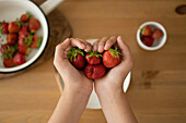 High angle of crop person demonstrating fresh strawberries in hands and in bowl with green leaves over wooden dining table indoors