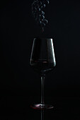 Eine stimmungsvolle Aufnahme, die ein Rinnsal Wein in ein Glas mit einer darüber schwebenden dunklen Weintraube vor einem schwarzen Hintergrund einfängt