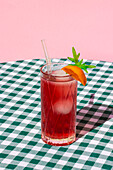 Glas kalter erfrischender roter Cocktail mit Eis und Strohhalm, serviert mit einer Orange auf einem Tisch mit kariertem Tischtuch
