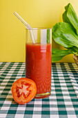 Transparent glass of fresh tomato juice with glass straw placed on checkered tablecloth against yellow background