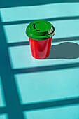 Minimalistischer roter Plastikbecher mit Kaffee zum Mitnehmen und grünem Deckel vor blauem Hintergrund, von oben gesehen