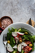 A vegetable salad made with fresh organic spinach leaves, sliced tomatoes, radishes, and olives presented in a white bowl beside a plaid cloth, wooden cutting board, and a bowl of pink salt