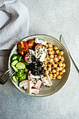 Top view of bowl with healthy salad and vegetables with chickpea sesame seeds and olives with cutlery and fabric placed on bright gray table