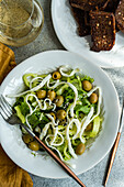 Draufsicht auf Gemüsesalat mit grünem Gemüse wie Kopfsalat, Gurken, Oliven, grüner Paprika mit hausgemachtem Käse und Sesam auf einem Teller mit Brot, einem Glas Wein und Besteck vor grauem Hintergrund