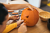 Anonymous women looking down while sitting at table and carving with tool scary teeth on orange pumpkin and preparing for Halloween decoration in day
