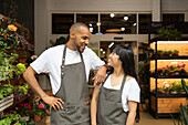 Cheerful young multiracial colleagues in aprons smiling and looking at each other while standing together in floristry store during work process