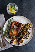 From above BBQ salmon steak seasoned with spices accompanied by fermented capers, charred lemon, a sprig of rosemary, with a refreshing lemon water on the side