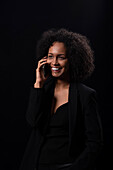 Positive Hispanic young female in outfit smiling and looking away while talking on mobile phone while standing on black background