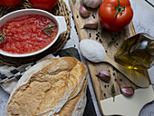 Draufsicht auf eine Schüssel mit Tomatenaufstrich und Vollkornbrot auf dem Tisch vor einer Flasche mit Olivenöl, Knoblauch und einem Löffel mit Salz