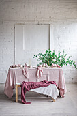 Wooden table with pink tablecloth and dishware placed near bench covered with cloth against potted plant and white brick wall