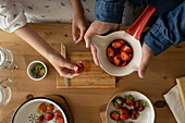 Top view of crop anonymous persons in casual clothes by wooden table with bowl and glass bottles while slicing strawberries and making jam indoors
