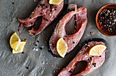 Top view of fresh trout steaks arranged on baking paper, ready to be cooked with Slices of lemon, salt and pepper near a small bowl of mixed fermented capers