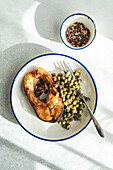 Top view of well-prepared grilled trout steak with capers served on a white plate with fork placed on grey surface near bowl with capers