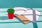 Minimalistischer roter Plastikbecher mit grünem Deckel, Notizbuch und Handy vor blauem Hintergrund von oben