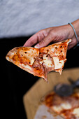 From above of anonymous person holding piece of delicious baked pizza with tomatoes in hand against blurred floor in daylight