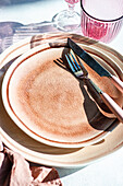 Ein elegant gedeckter Tisch mit strukturierten Keramiktellern, modernem Besteck und rosa Gläsern, die im Sonnenlicht weiche Schatten werfen