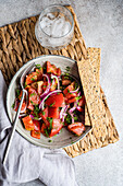 Schale mit Salat aus Tomaten, roten Zwiebeln und Dillkräutern in der Draufsicht neben einem mit Eis gefüllten Glas
