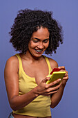 Cheerful Hispanic female with Afro hairstyle in yellow top holding credit card and browsing smartphone while standing on blue background