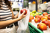 Anonyme Kundin in Freizeitkleidung wählt frische Äpfel aus und packt sie in eine Plastiktüte auf dem Markt